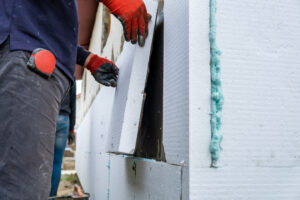 Construction worker installing styrofoam insulation sheets on house facade wall for thermal protection.